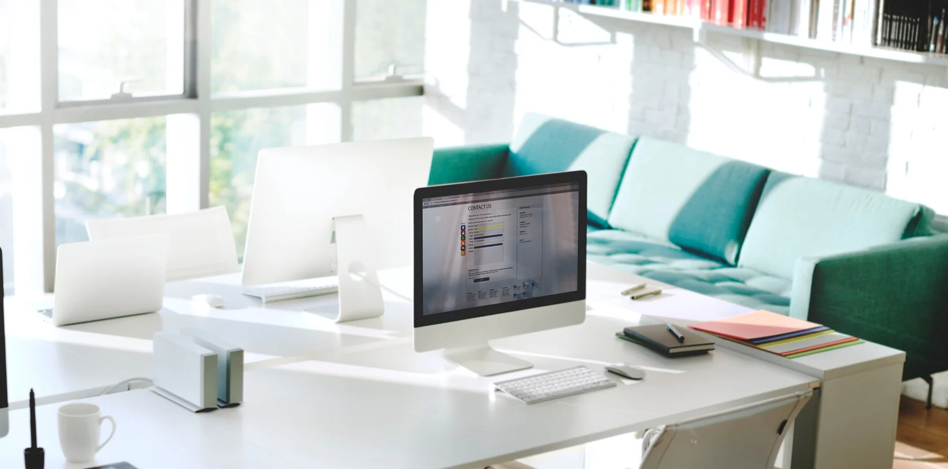 Bright modern office with a white desk, iMac monitor, keyboard, and colorful notebooks; large windows flood the space with sunlight and a teal sofa in the background.